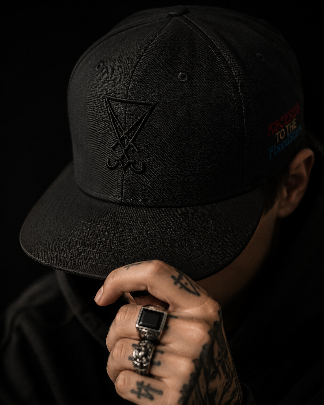 Person wearing a black cap with a symbol, holding a watch with tattoos on the hand, against a dark background.