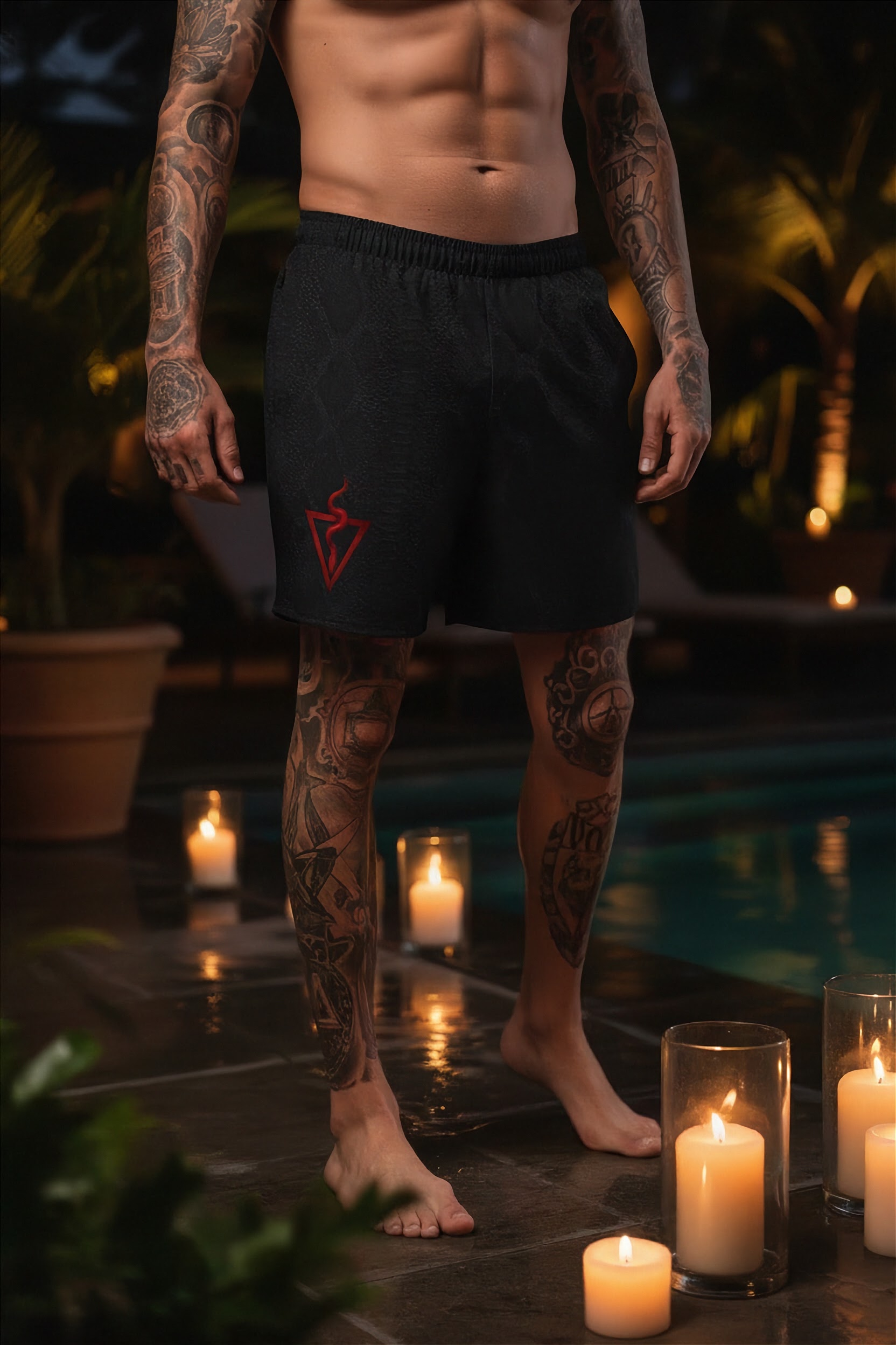 Man with tattoos wearing black shorts with a red logo by candles by a pool